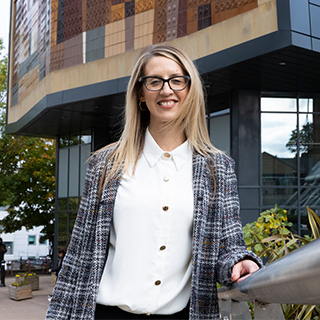 A student wearing glasses, a white shirt, and a checkered jacket holds the railings outside the university campus building while smiling at the camera.