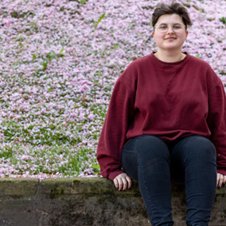 Student wearing glasses sat on a wall smiling with blossom in the background.