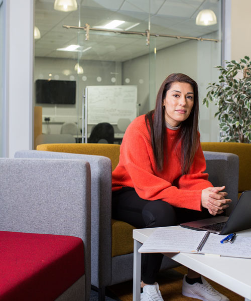 A smartly dressed person sitting in an office environment and looking at the camera.