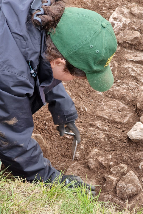 A person with a green cap and a raincoat digs in the earth with their back to camera.