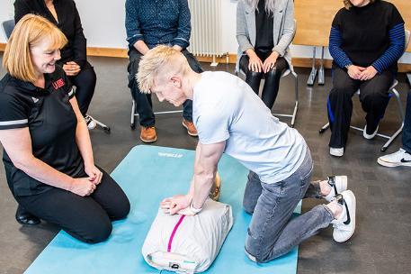 A First Aid student performs a resuscitation technique on an adult mannequin in front of the First Aid instructor whilst other students look onlooks