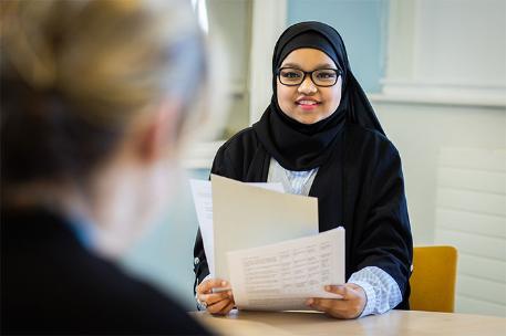 Two people sat at a table. One of them is holding some documents.