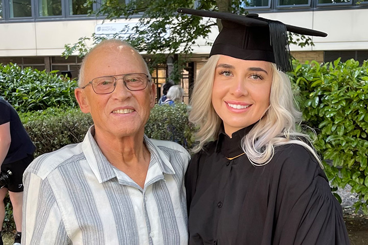 A student wearing graduation gown smiling at the camera alongside a senior family member who is also smiling at the camera