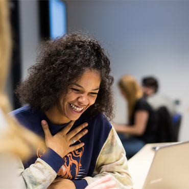 A student smiling while studying on a laptop in a shared study space on campus.