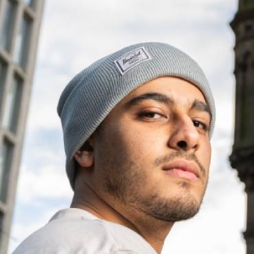 A man in a grey hat stands in front of tall buildings.