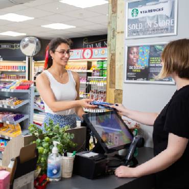 A woman in a white vest speaks to a different woman stood behind the counter of a shop.