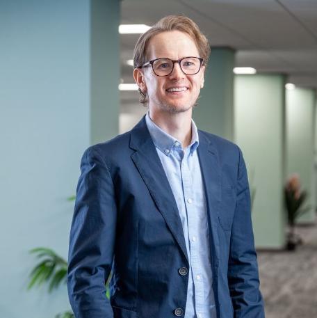 Alumnus Ben Foster, wearing glasses and a blue shirt in a modern office environment.