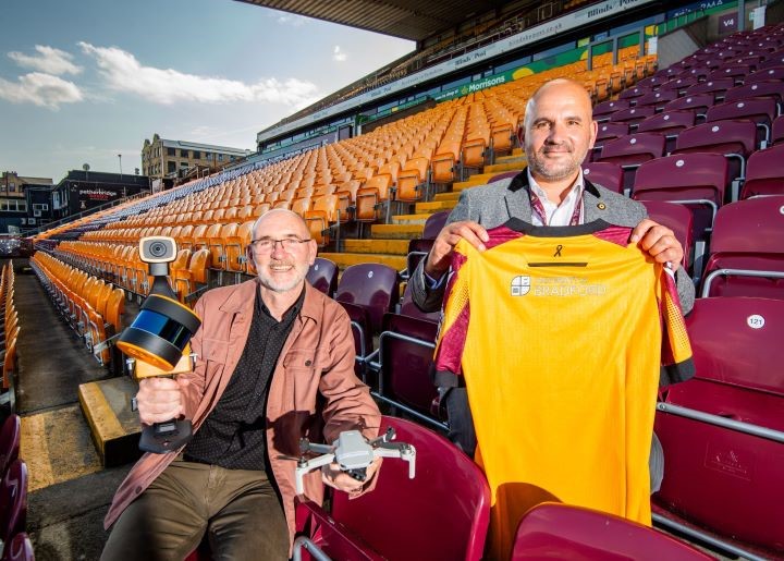 Chris Gaffney, Professor of Archaeology, and Davide Longo, Chief Commercial Officer of Bradford City Association Football Club, sat in the stands at the University of Bradford stadium. Chris Gaffney is holding a drone and a 360 camera while Davide Longo is holding up a Bradford City Association Football Shirt