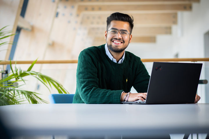 A person sitting at a table with a laptop, smiling at the camera.