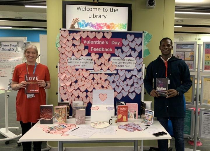 A member of staff and student standing in front of a notice board with heart shaped post-it notes on it