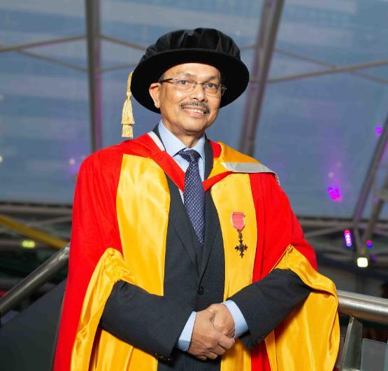 Honorary graduate in gown stands next to barrier smiling