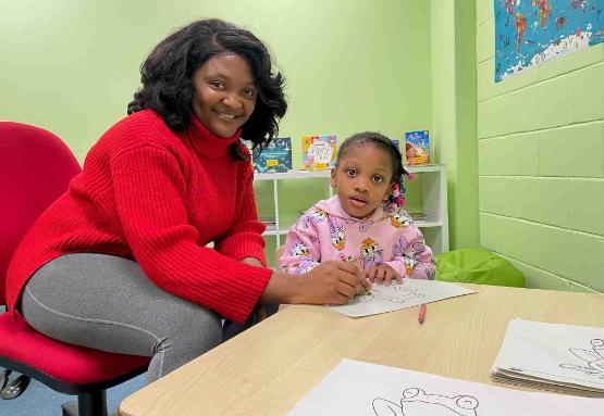 Student sat at small table with young child who is colouring in a piece of paper