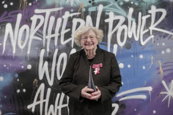 An academic holds an award stood in front of banner with large graffiti style lettering written on it