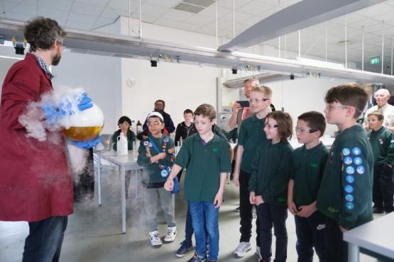 A scientist shows off a large glass container which is bubbling over to a group of people in a university room