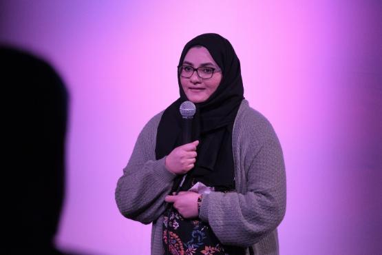 A person wearing a head dress holds a microphone and delivers a stand-up comedy routine