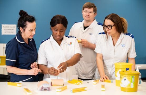 A group of student nurses in uniform stand up behind a table with a number of drugs on it that they are working with