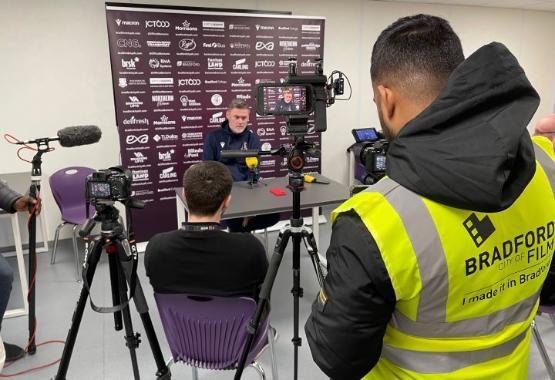 a student stands behind video camera on a tripod filming a press conference with a manager sitting at a desk