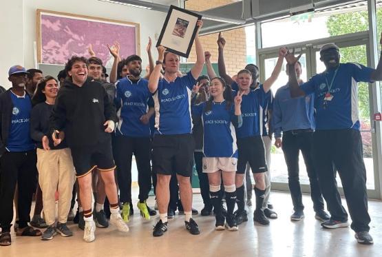 a large group of students and staff in football kit celebrate with a trophy