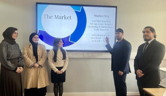 THree students stood on the left and another two students stood on the right present a pitch using a projector screen on a wall behind them