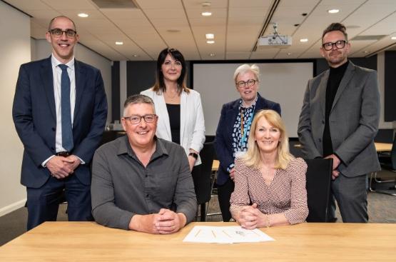 Two people sat behind a wooden table with four other people stood behind them smiling at signing event