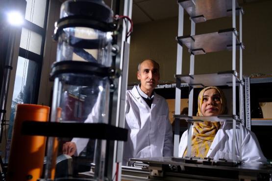 A university staff member and student in white coats standing next to each other in a laboratory looking at a structure of small metal shelves