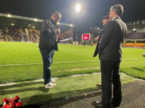 A person holding a microphone stands to the side of two university staff members who are being interviewed on the touchline at a football stadium