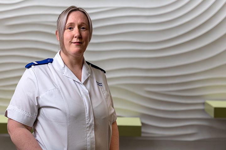 A student in a nursing uniform stood in front of a wave textured background. They are smiling at the camera.