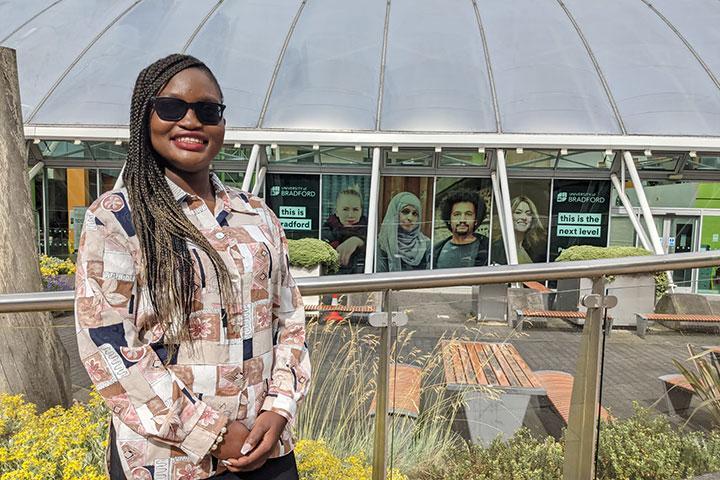 A person standing outside wearing sunglasses, in front of a window display advertising postgraduate study at Bradford.