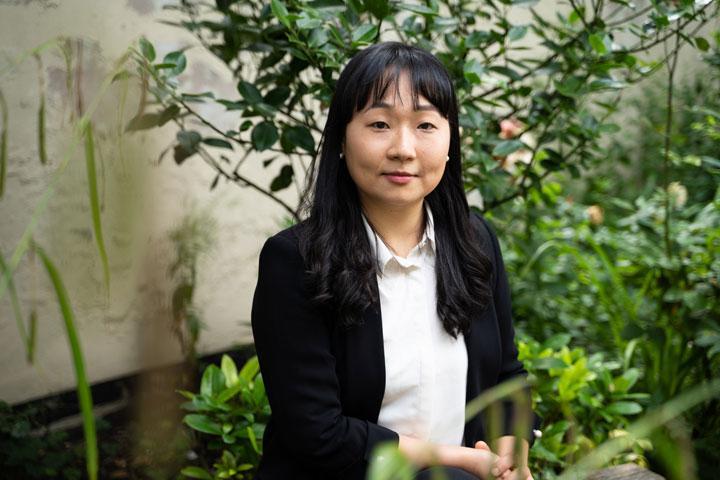 A smartly dressed person sitting down and smiling at the camera, with plants in the background.