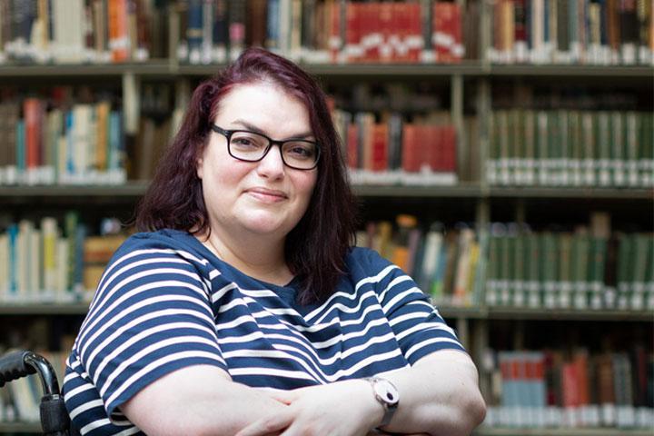 A head and shoulders image of a person in a library, smiling at the camera with arms folded.