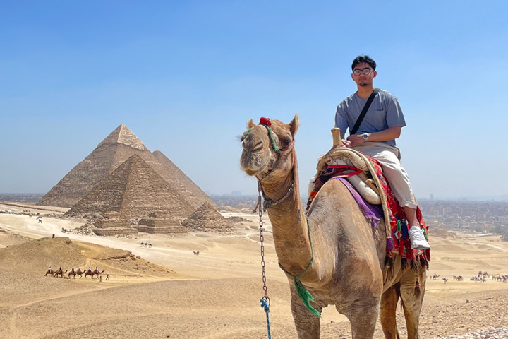 A student riding a camel, posing in-front of the Great Pyramids in Egypt