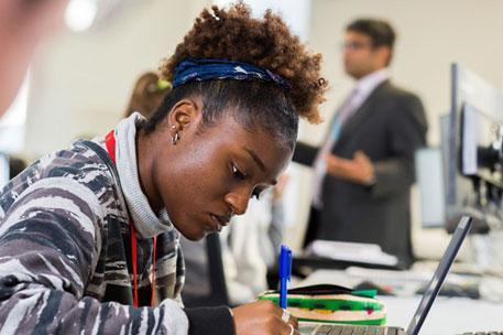 A student sitting at a desk writing with a lecturer in the background