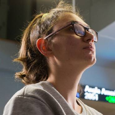 A student with glasses and long hair looks to the right of the camera with bright light above them.