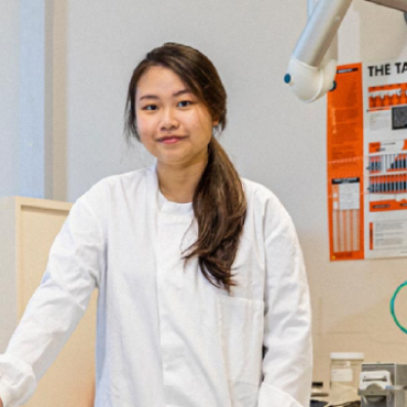 A student in a white uniform stands next to medical equipment while looking into the camera with a faint smile.