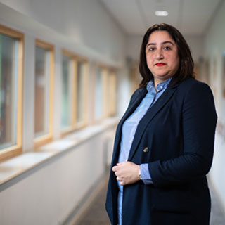 A smartly dressed person standing, facing the camera in a corridor in a health setting.